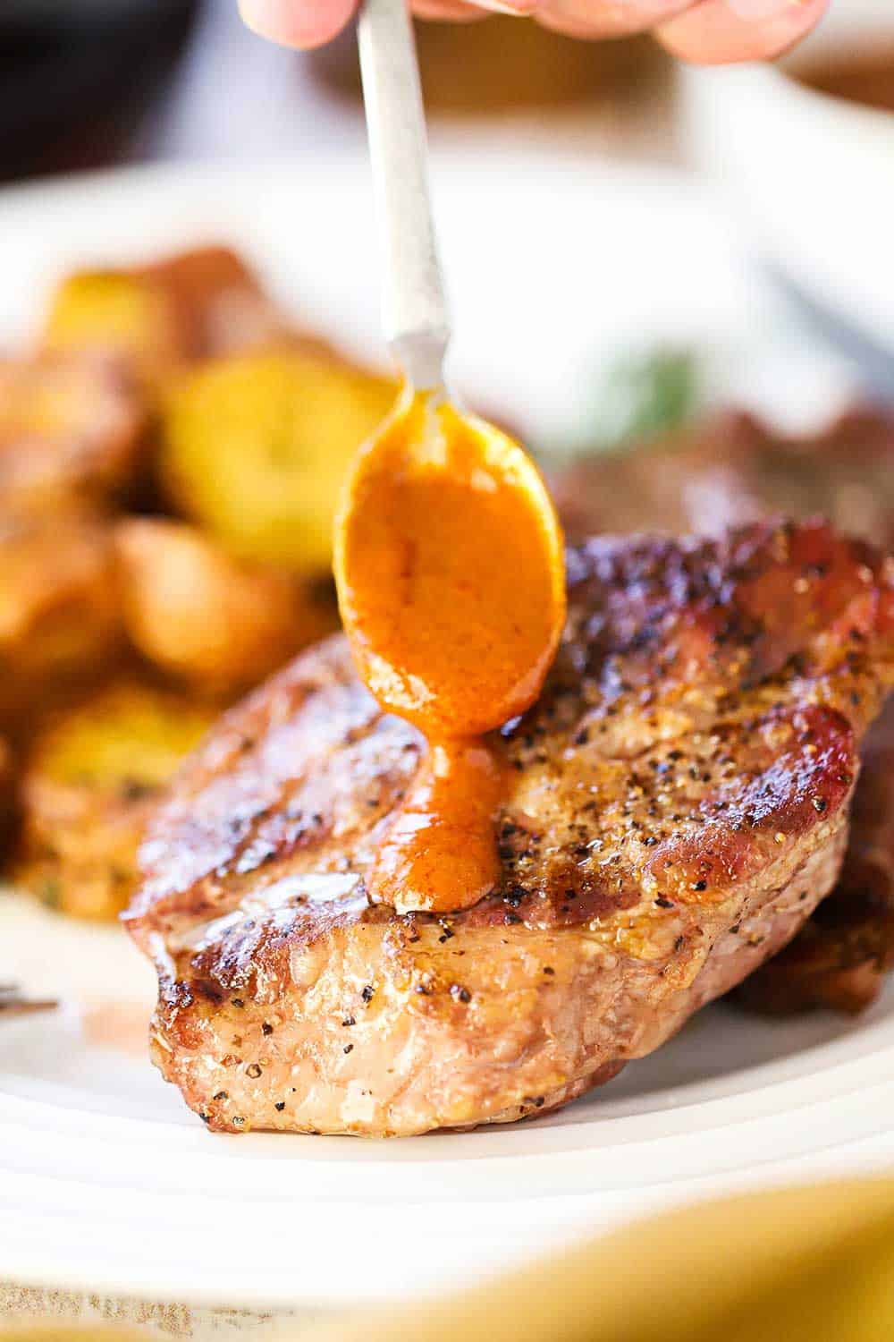 A hand holding a small spoon with steak sauce on it drizzling over a pork steak on a white plate next to roasted potatoes.