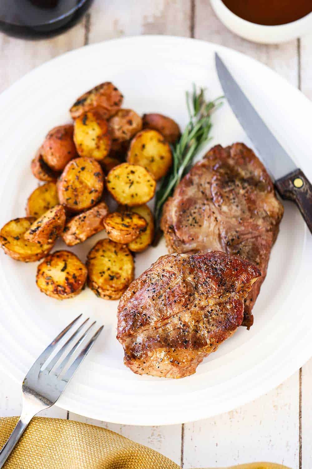 An overhead view of a white dinner plate with a grilled Delmonico pork steak on it next to a sprig of rosemary and roasted new potatoes.