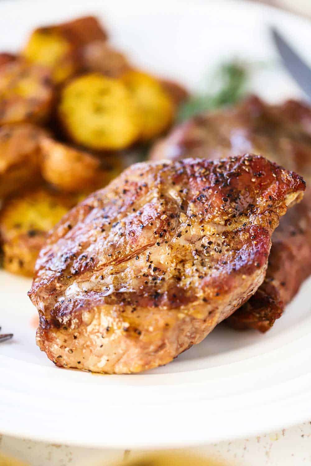 A close up view of a grilled Delmonico pork steak on a white plate next to roasted potatoes.