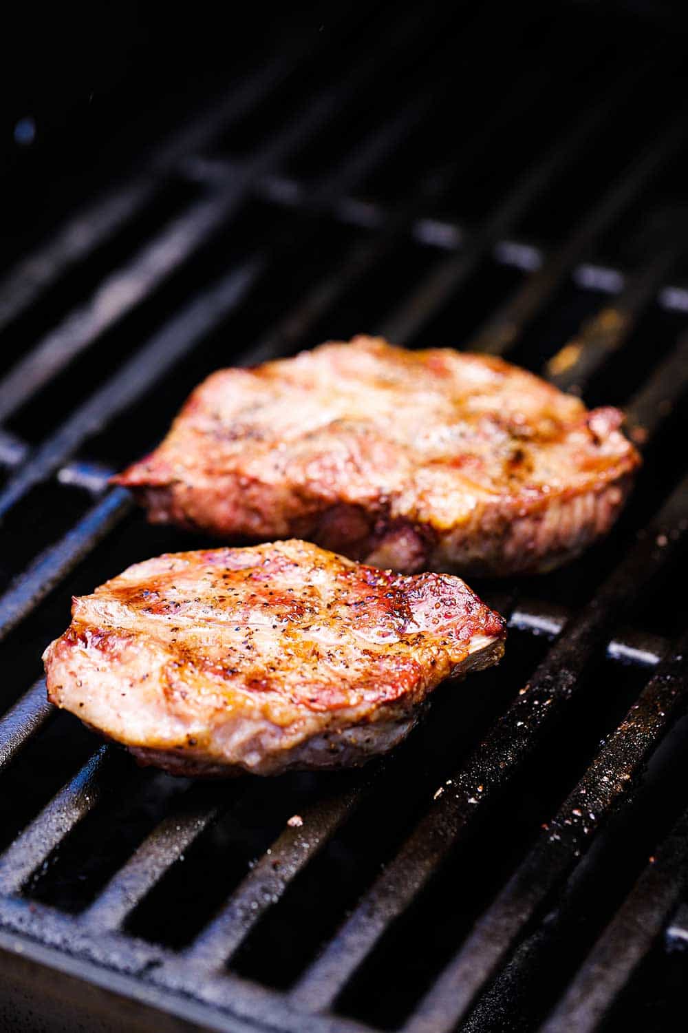 Two pork steaks on a gas grill seasoned with salt and pepper.