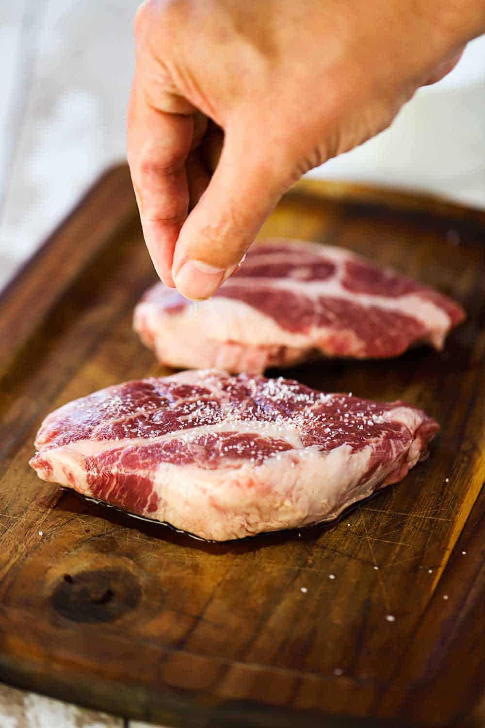 A hand sprinkling salt on a pork steak on a small wooden cutting board next to a second pork steak.