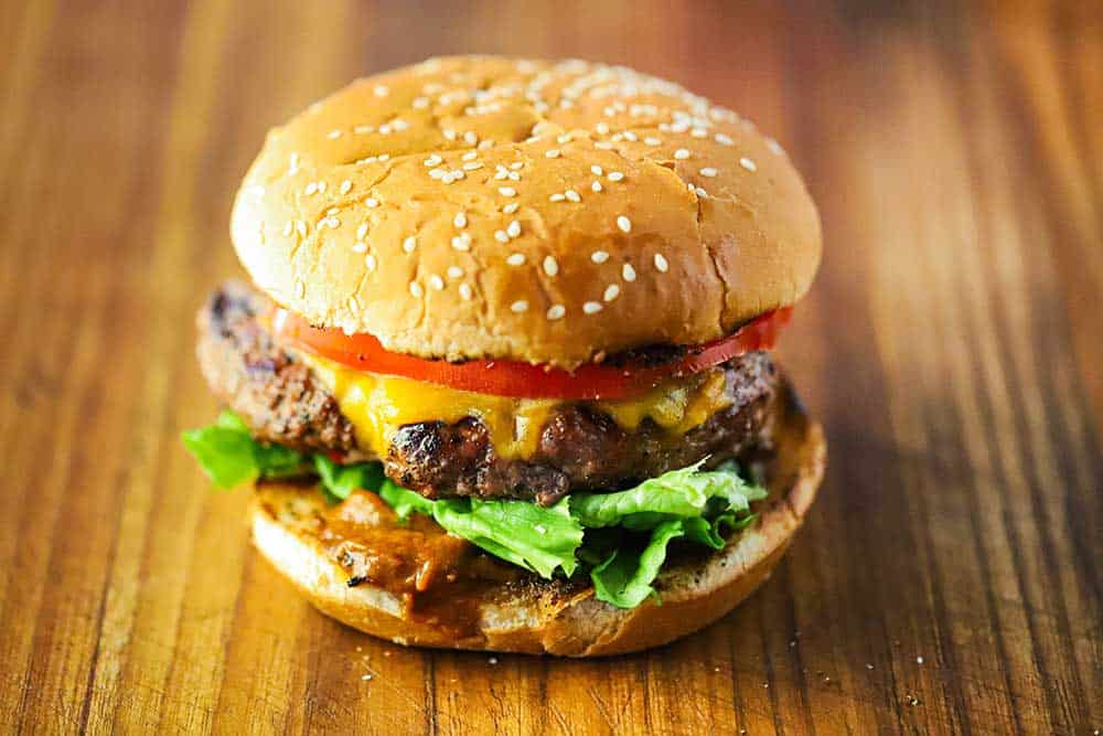 A pork burger with a sliced of green lettuce under the patty and a slice of tomato on top all on a wooden cutting board.