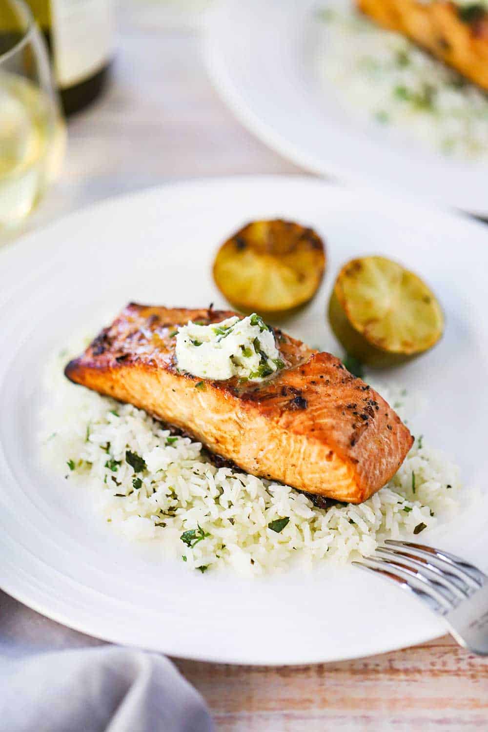 A grilled salmon fillet sitting on top of a bed of cilantro rice with poblano compound butter melting on top of the dish with two grilled limes halves nearby.