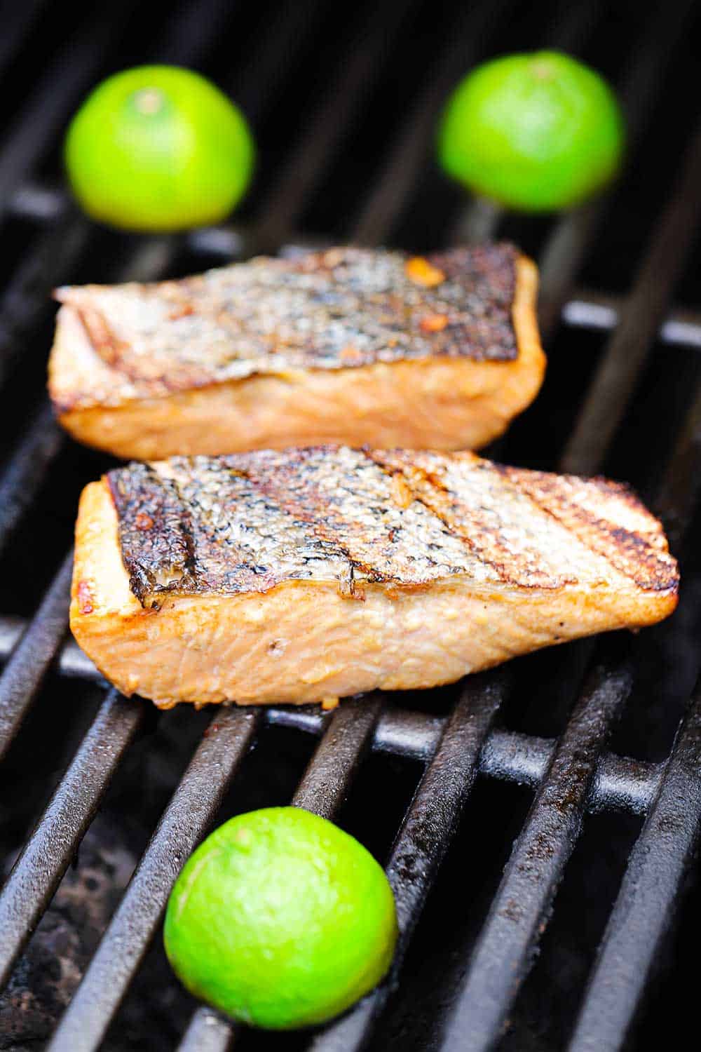 Two salmon fillets on a gas grill with three lime halve on the grill, cut-side down.