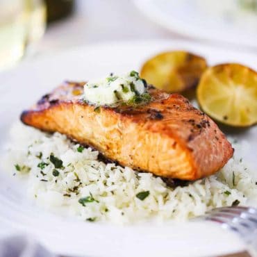 A close up view of a fillet of grilled salmon topped with a pad of melting poblano butter sitting on a bed of cilantro rice on a white plate.