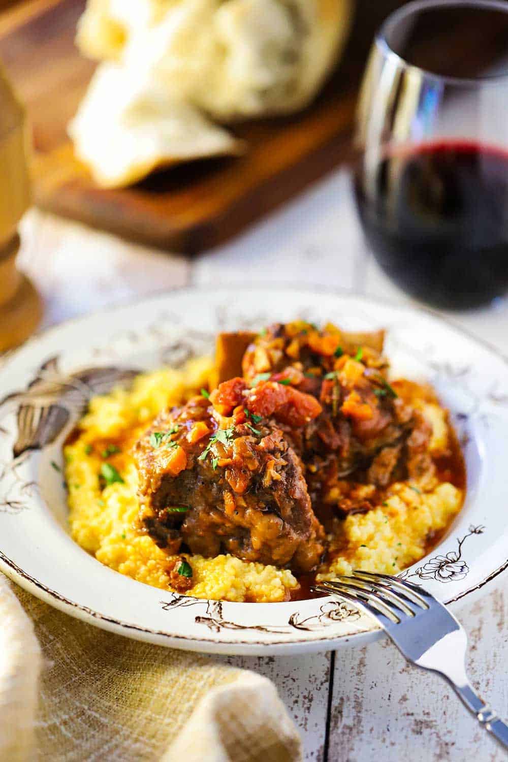 An antique bowl filled with polenta and topped with braised Italian short ribs next to a glass of red wine. 