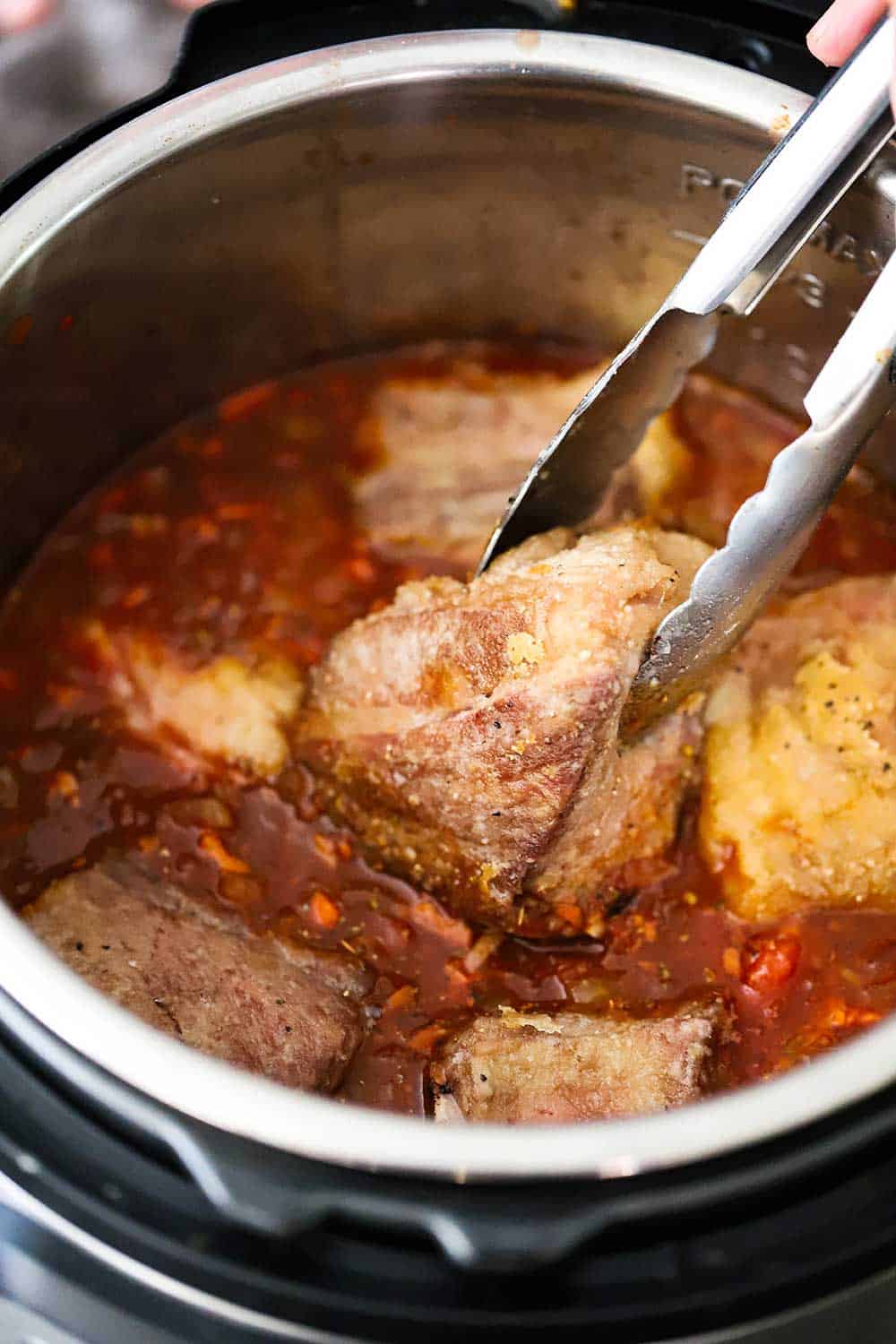 A pair of tongs lowering seared beef short ribs into an Instant Pot filled with a wine and beef broth sauce. 