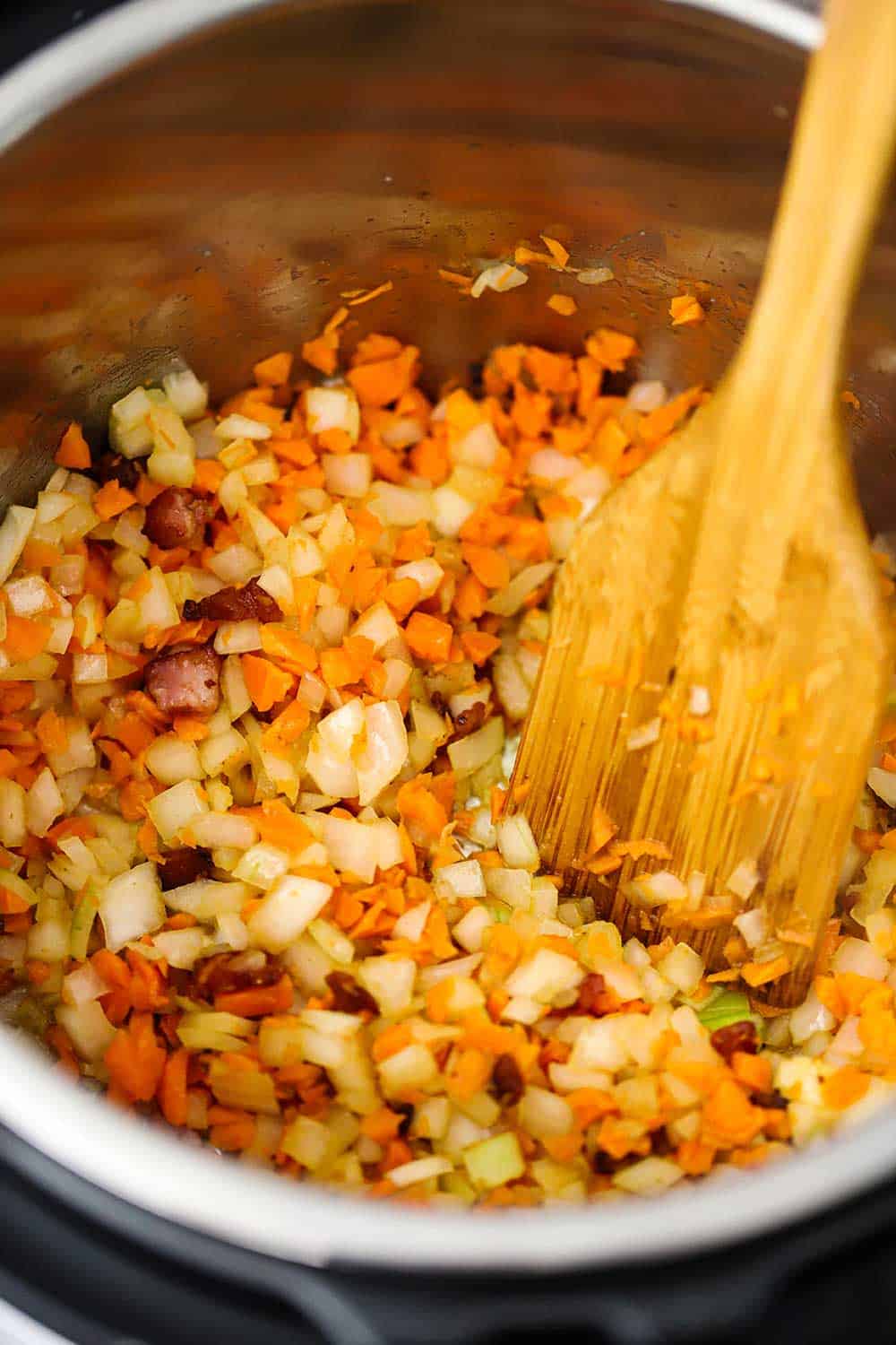 An overhead view of sautéed onions, carrots, and pancetta in an Instant Pot with a wooden spoon in it. 