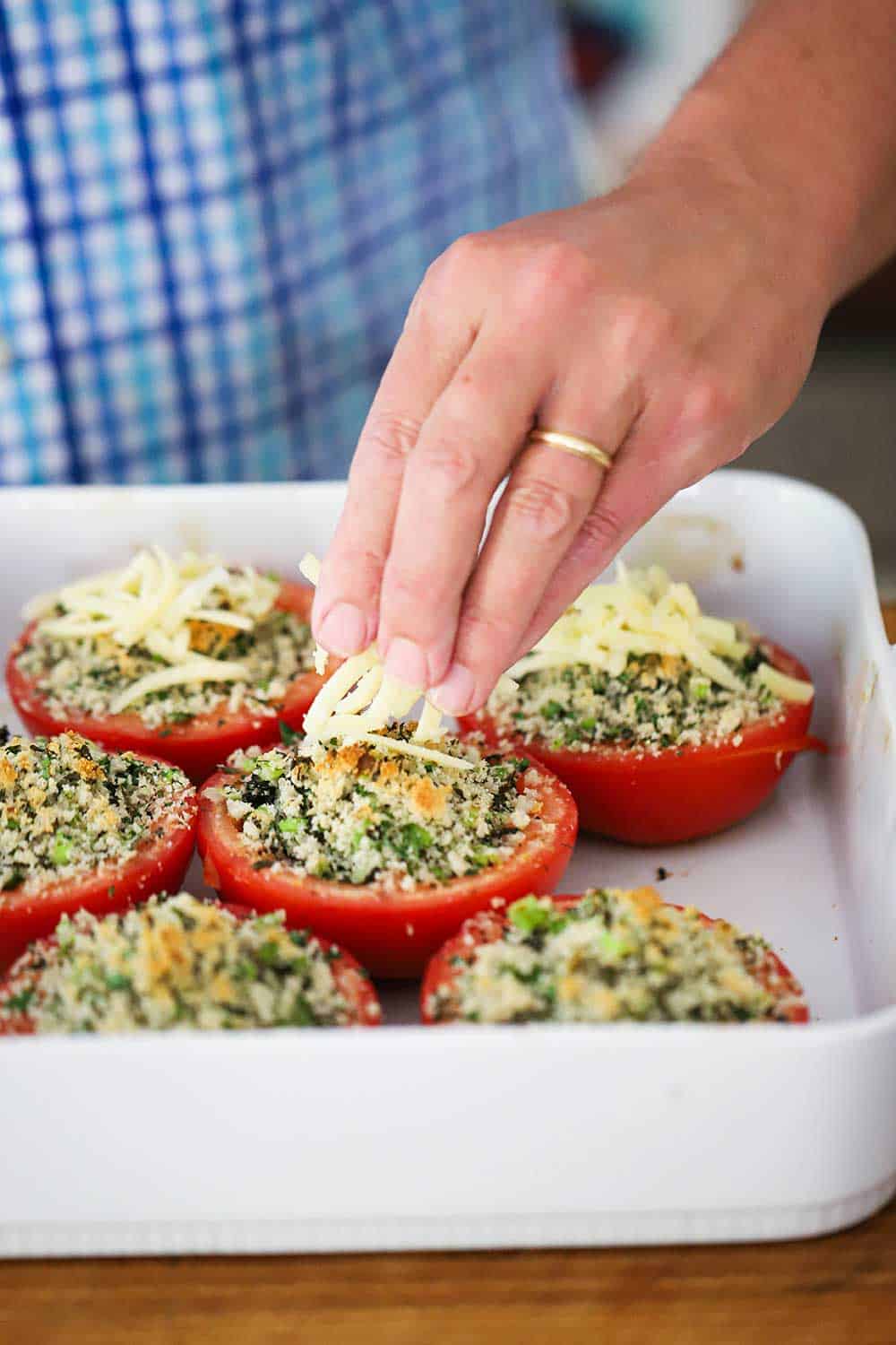 A hand sprinkling shredded cheese over roasted halved stuffed tomatoes in a white baking dish.