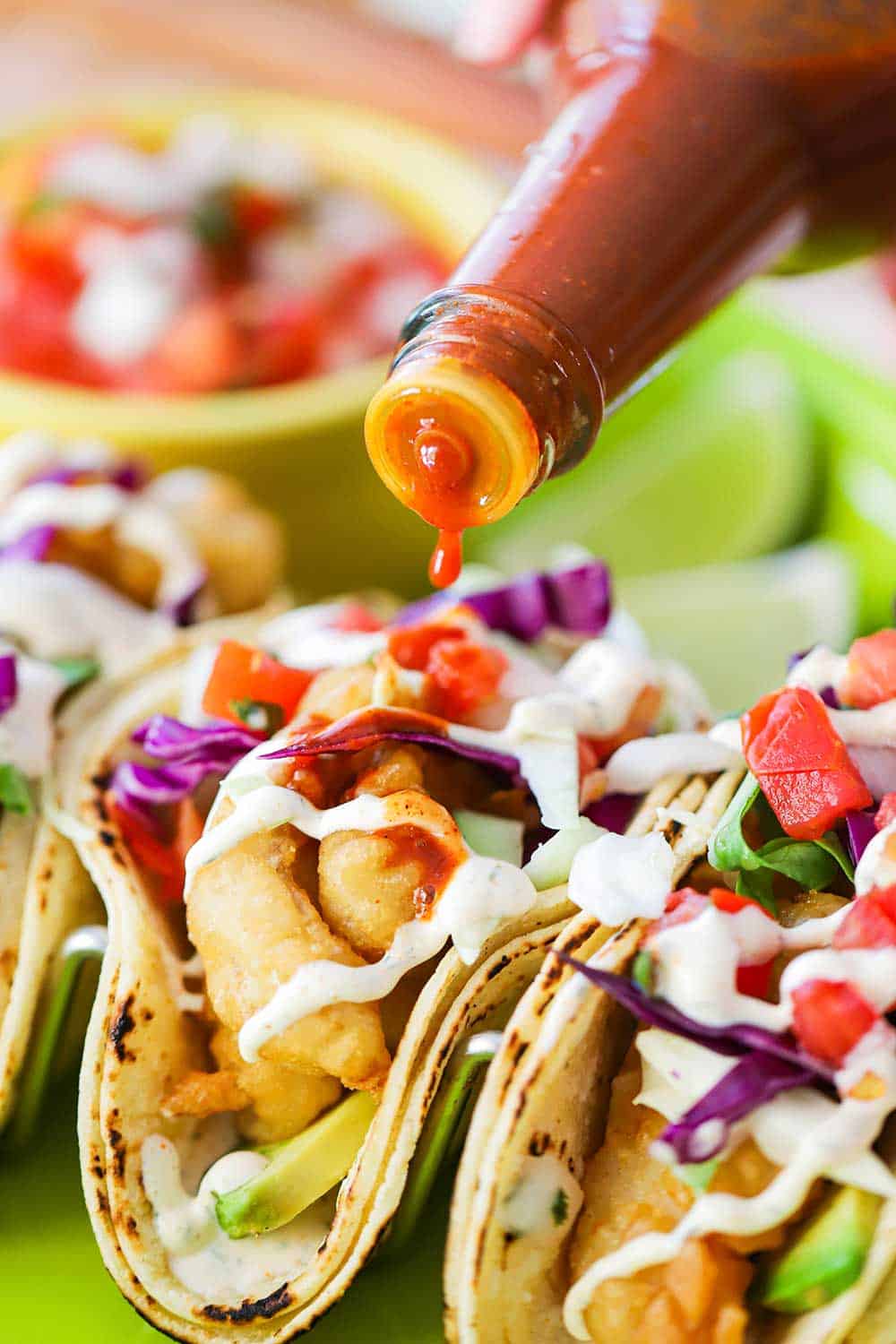 A close up view of a bottle of hot sauce drizzling sauce over Baja fish tacos.