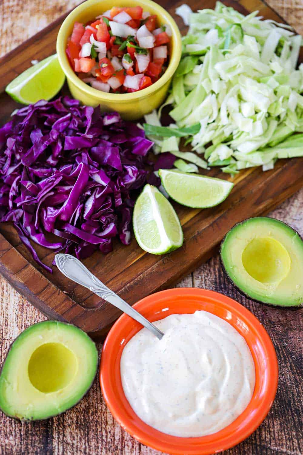 An overhead view of a bowl of Baja sauce, sliced avocados, chopped cabbage, lime wedges, and pico de gallo.