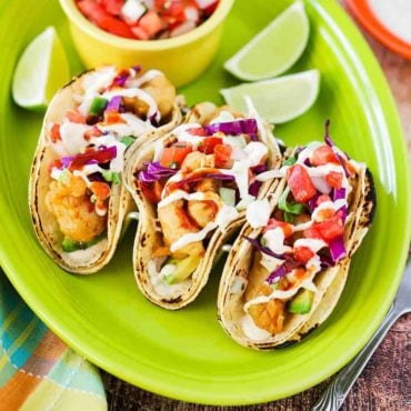 An overhead view of three fish tacos sitting on a green platter next to lime wedges and a bowl of pico de gallo.