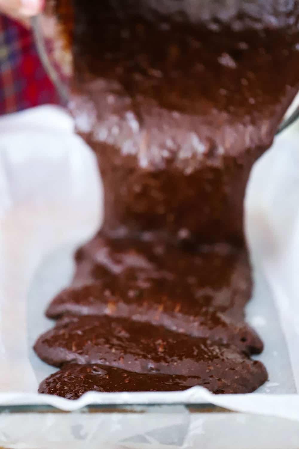 Fudge brownie batter being poured into a baking dish lined with parchment paper. 