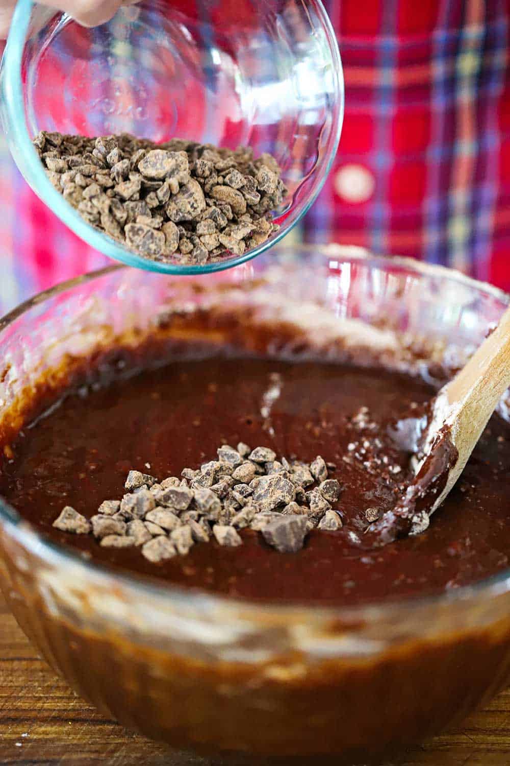 A small bowl of chopped semi-sweet chocolate being dumped from a small bowl into a large bowl of brownie batter. 