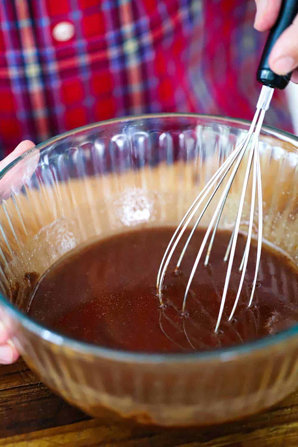 A glass bowl filled with melted chocolate and butter with a hand whisking the mixture with a wire whisk. 