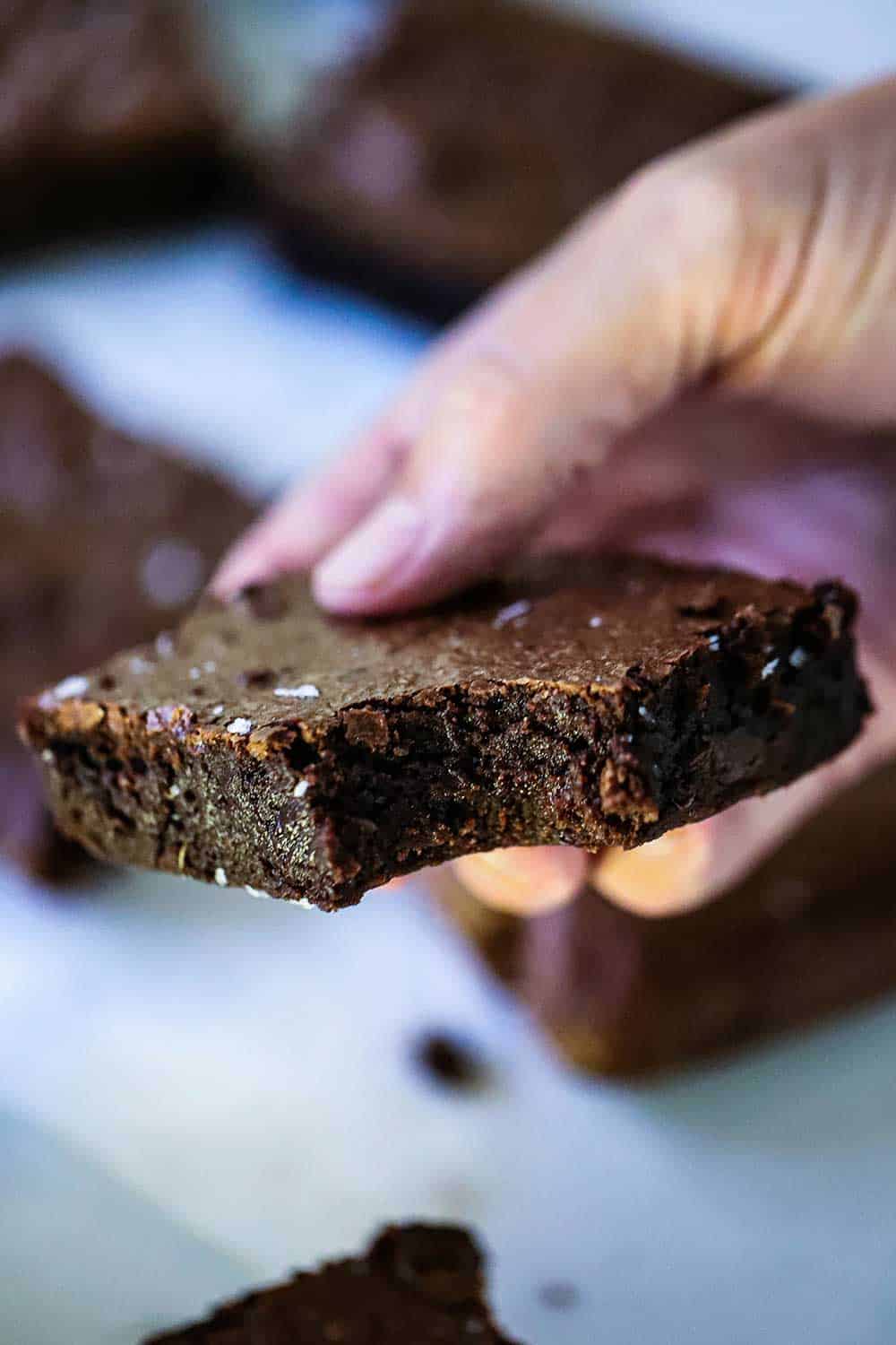 A hand holding a fudge brownie with a bite taken out of it.