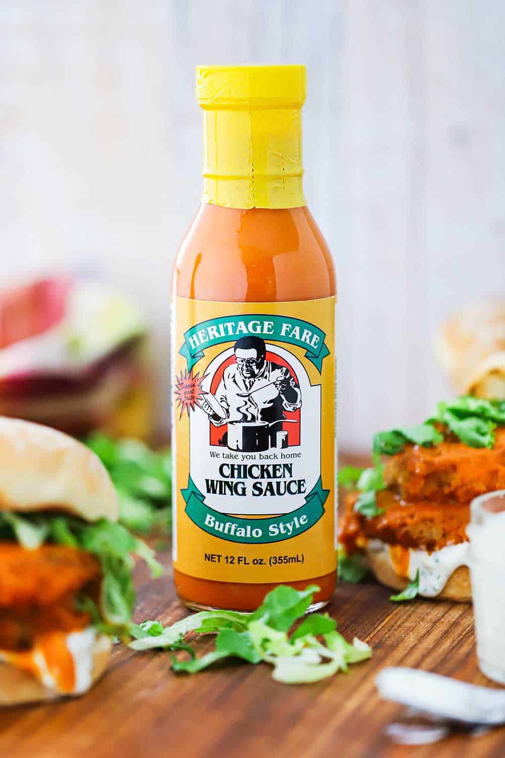 A bottle of Heritage Fare Buffalo Chicken Wing sauce next to a buffalo chicken sandwich on a cutting board. 