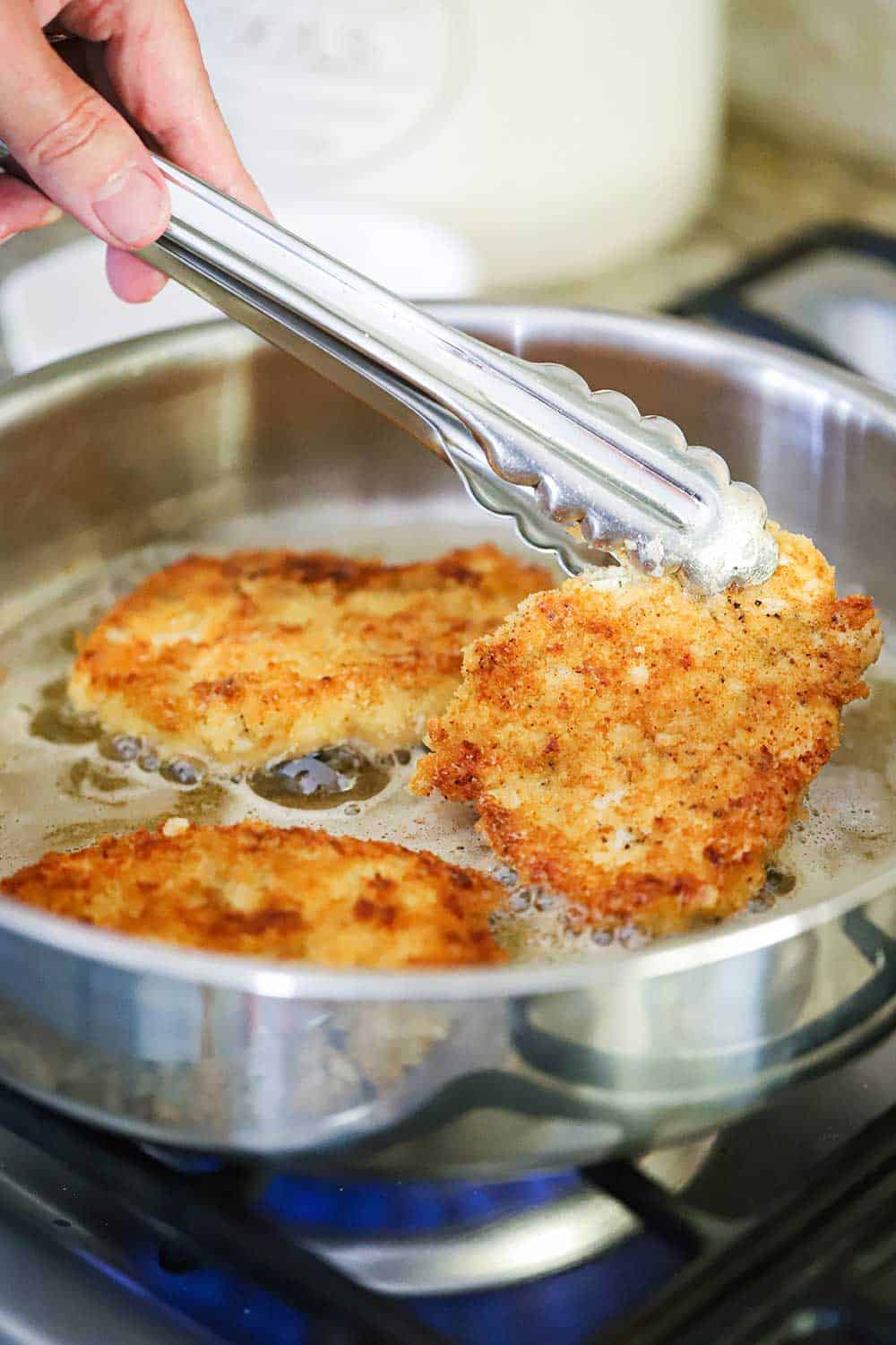 Three chicken cutlets frying in a large saucepan in oil and butter with tongs holding one up. 