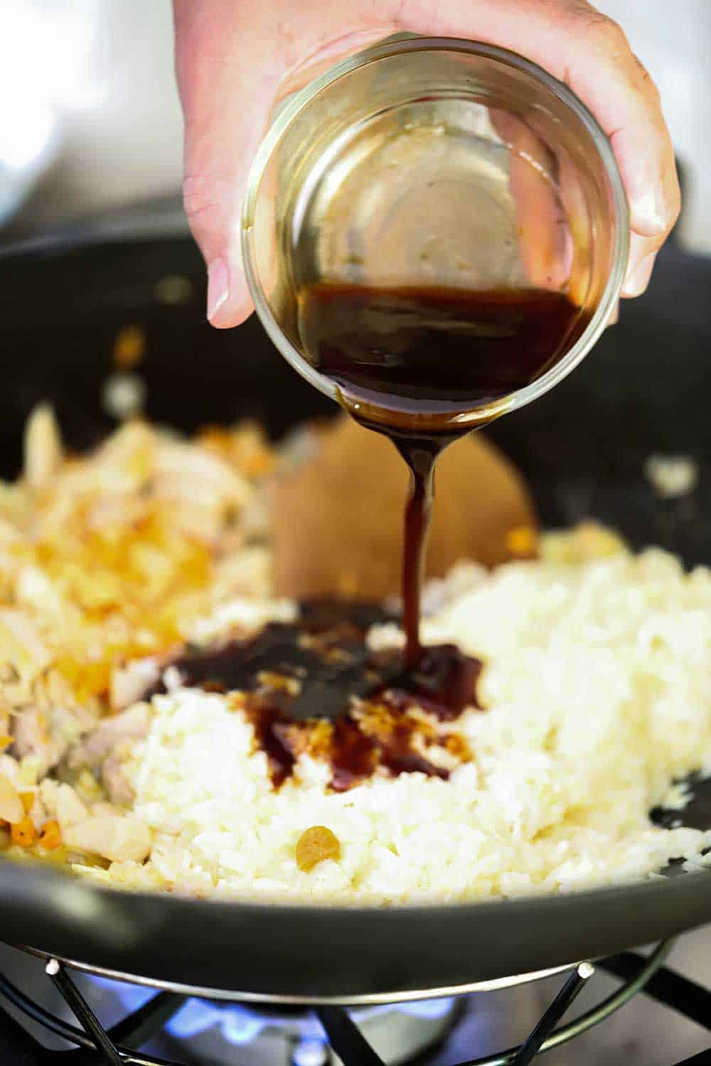 A hand pouring oyster sauce from a small glass bowl into a wok filled with fried rice. 