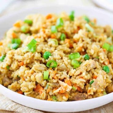 A close-up view of a white serving bowl filled with homemade chicken fried rice next to a gold spoon.