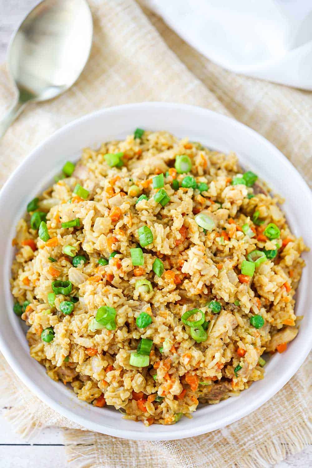 An overhead view a white serving bowl filled with chicken fried rice next to a gold spoon. 