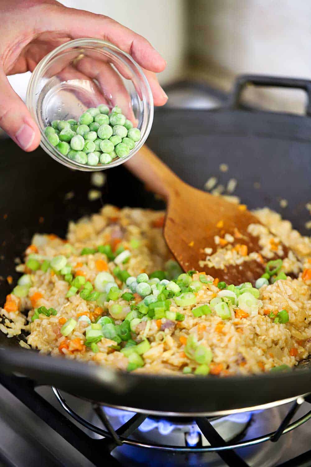 A hand dropping frozen peas into a wok filled with chicken fried rice and a wooden spoon. 