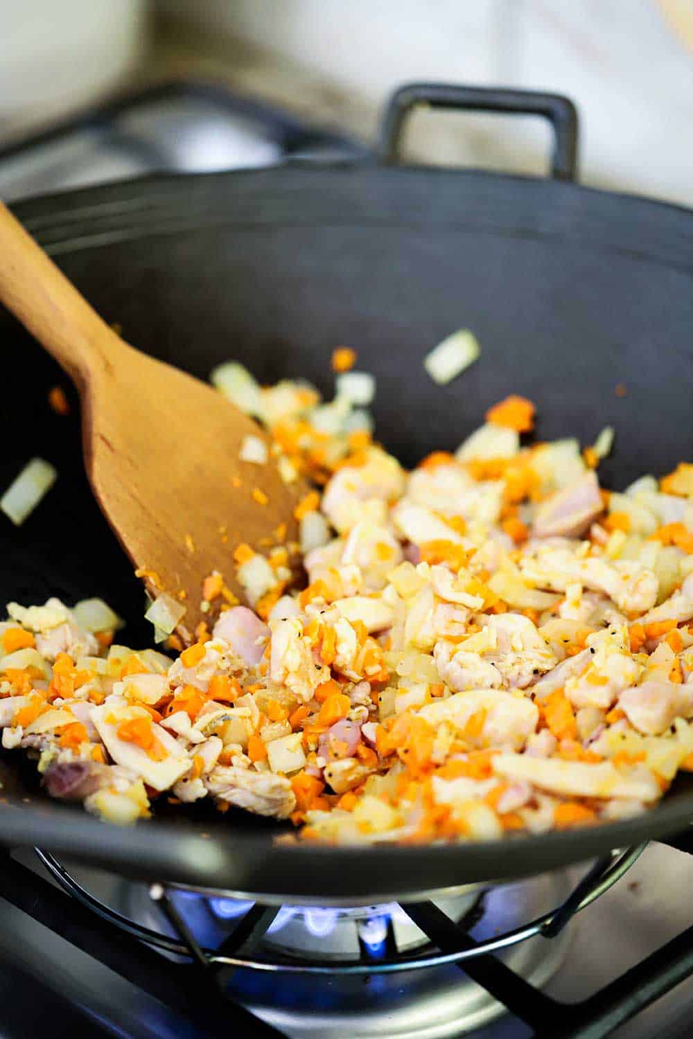 A metal wok over a gas stove with satuéed onions, carrots, and chicken in it, with a wooden spoon stirring it all. 