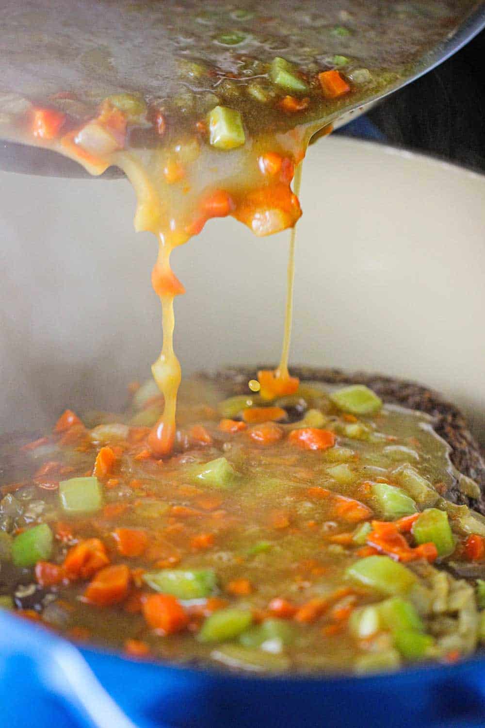 Sauteed vegetables and broth being poured of a roast in a Dutch oven. 