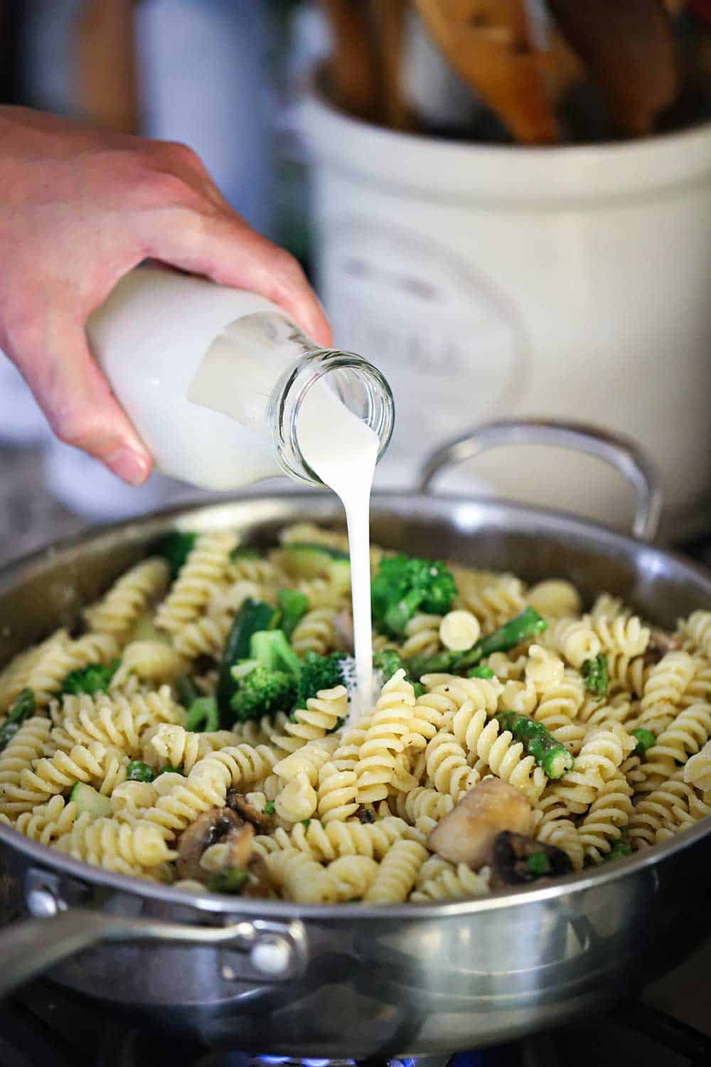 A hand pouring cream from a milk jar into a skillet filled cooked pasta and broccoli.