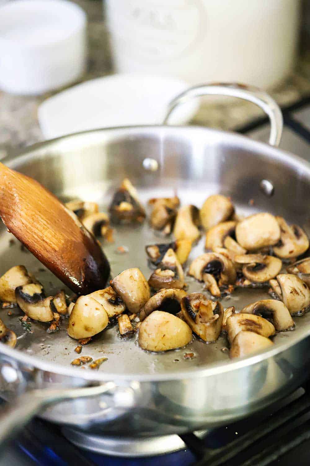 A large stainless steel skilled filled with quartered button mushrooms and garlic, all being sauteed.