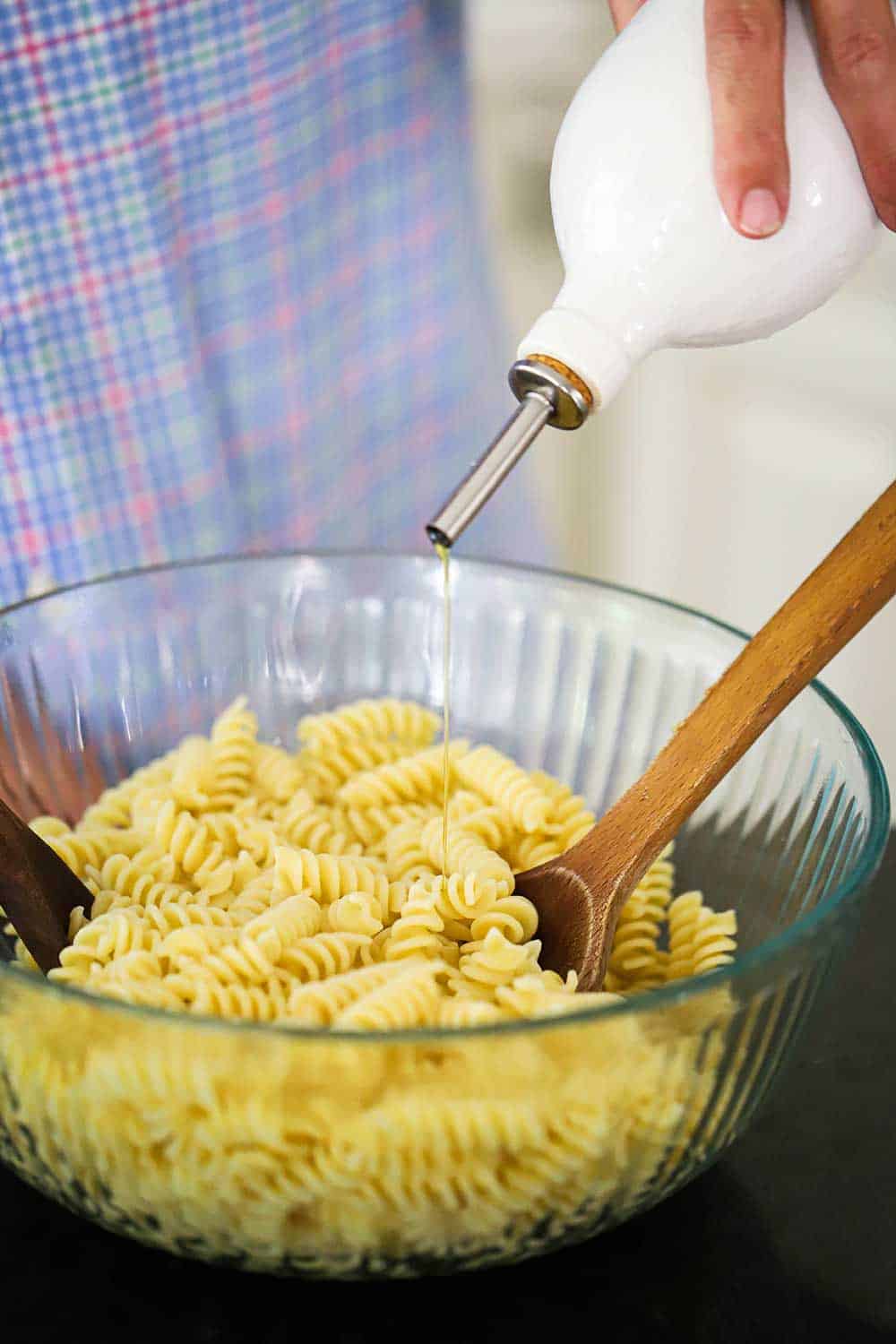 A large glass bowl filled with cooked pasta and a person pouring olive oil into it from a dispenser.