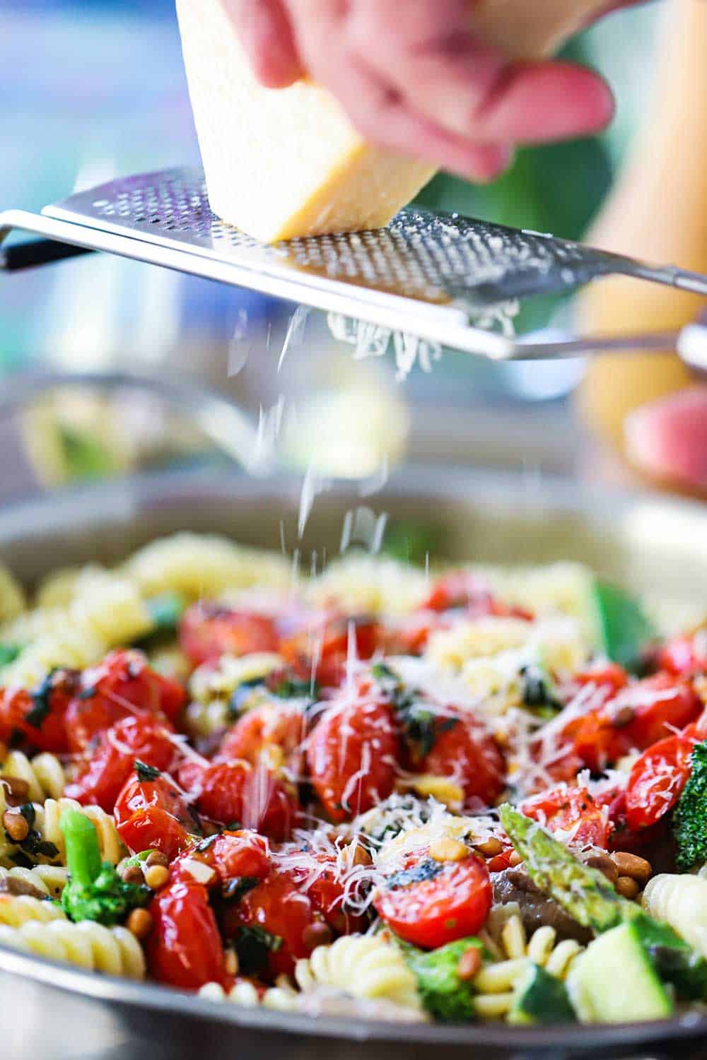 A hand scraping a block of Parmesan cheese over a microplane with grated cheese falling into a skillet of pasta primavera.