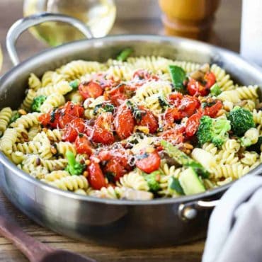 A large stainless steel skilled filled with pasta primavera topped with sauteed tomatoes and shaved cheese.