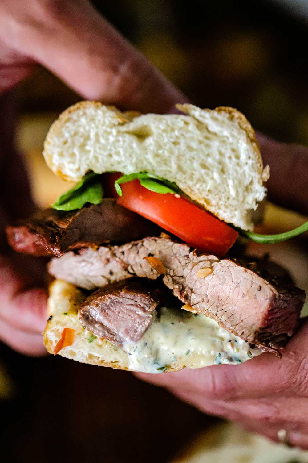 A close-up view of a grilled steak sandwich that is cut in half and is being held a pair of hands. 