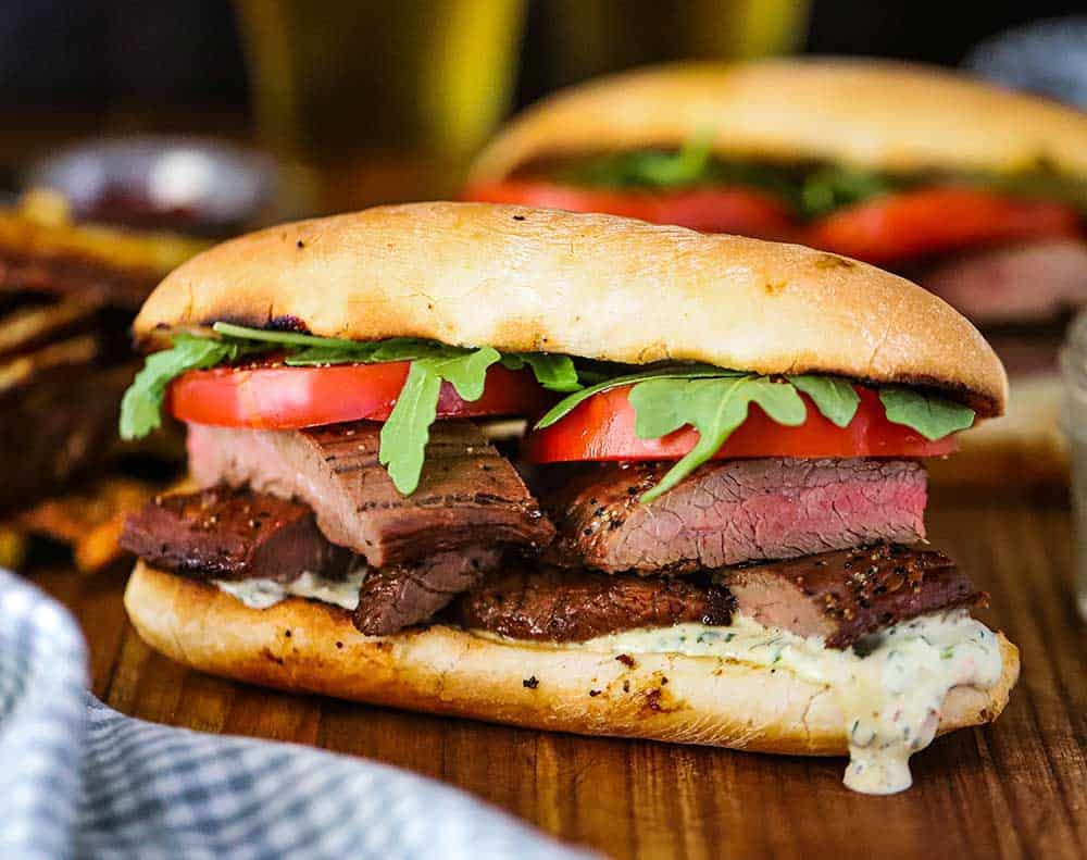 A grilled steak sandwich loaded with chimichurri mayonnaise, layers of juicy steak, and topped with tomatoes and arugula, all on a hoagie. 