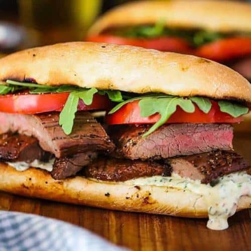 A grilled steak sandwich loaded with chimichurri mayonnaise, layers of juicy steak, and topped with tomatoes and arugula, all on a hoagie.