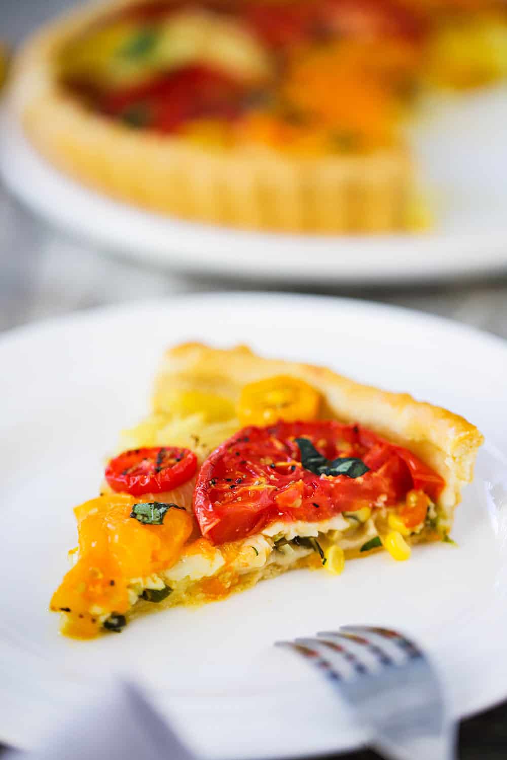 A slice of a tomato tart with corn and herbs on a small white plate next to a fork.