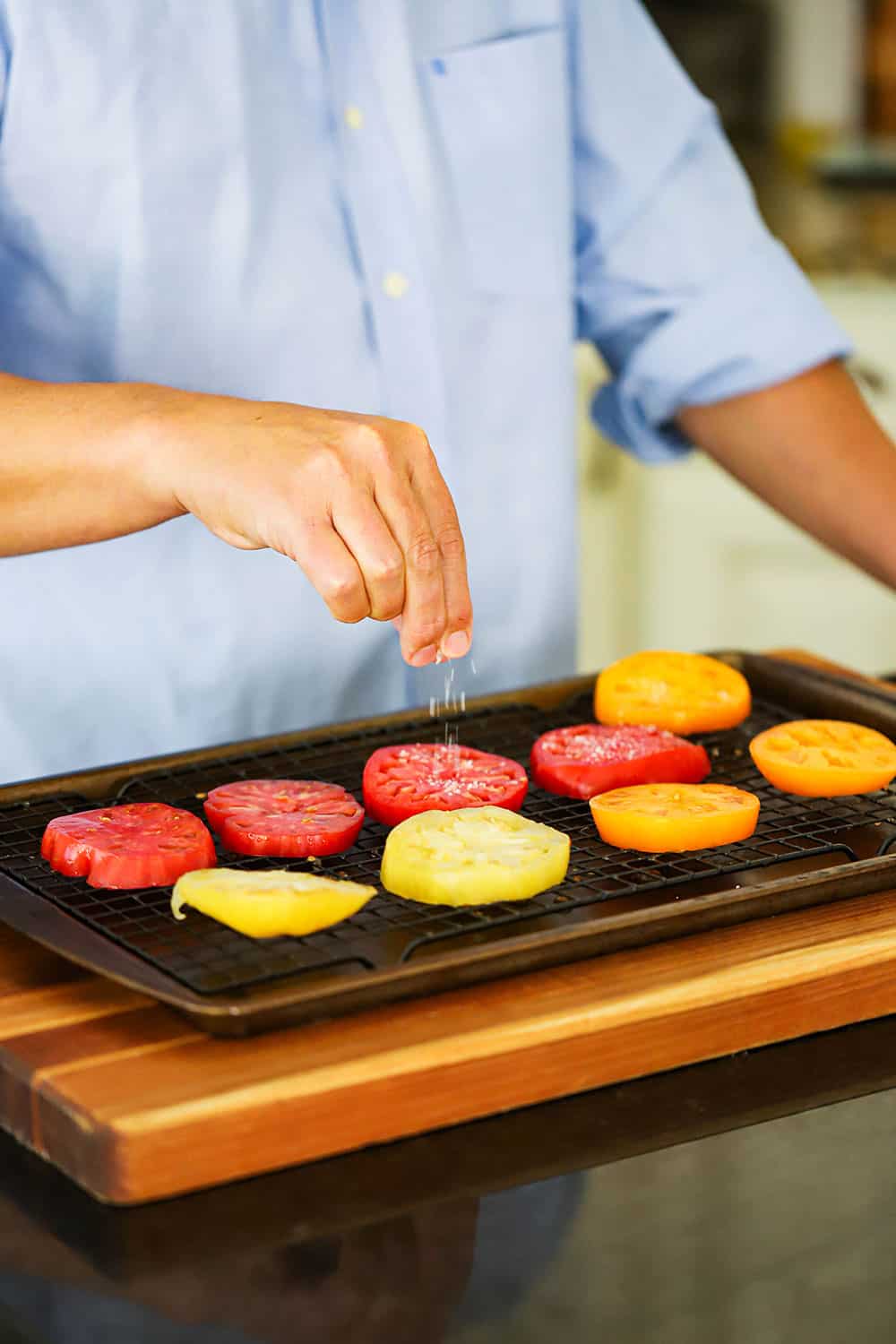 A person sprinkling salt onto sliced tomatoes on a baking rack in a baking sheet on a cutting board. 