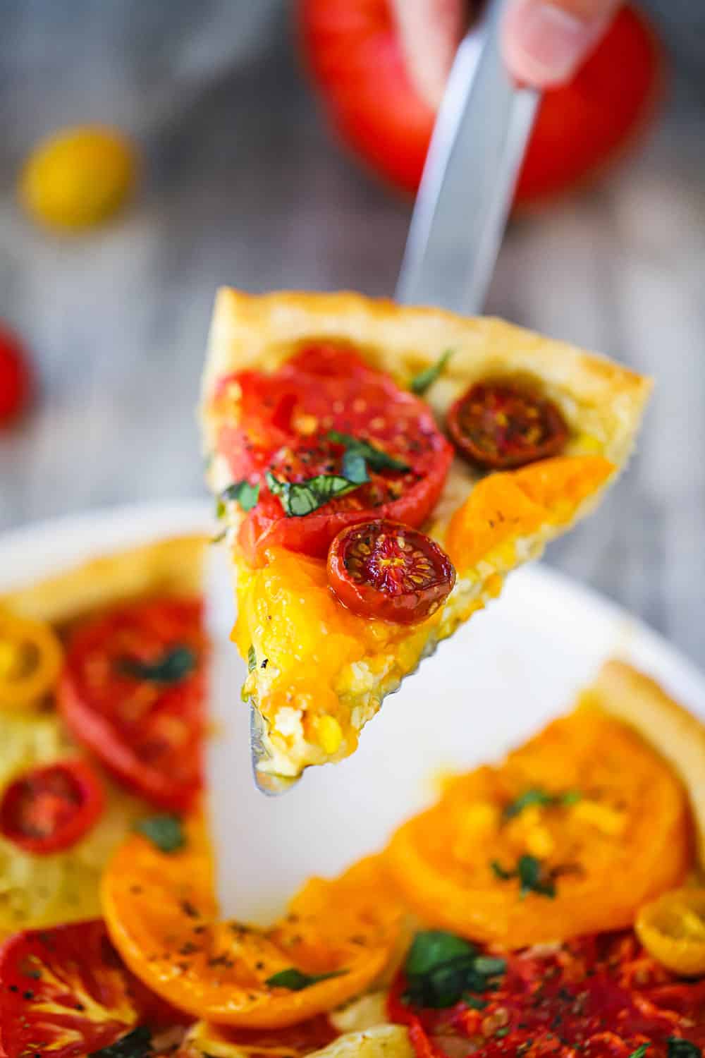 A spatula holding up a slice of tomato tart with corn from a serving plate. 