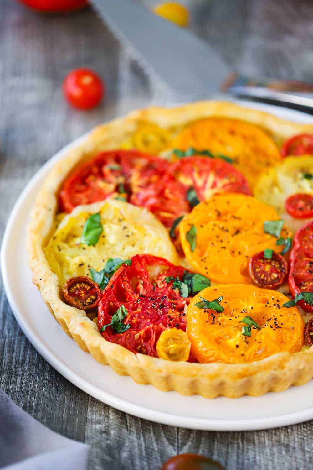 A tomato tart with colorful sliced tomatoes on top in a pastry shell all on a white serving plate. 