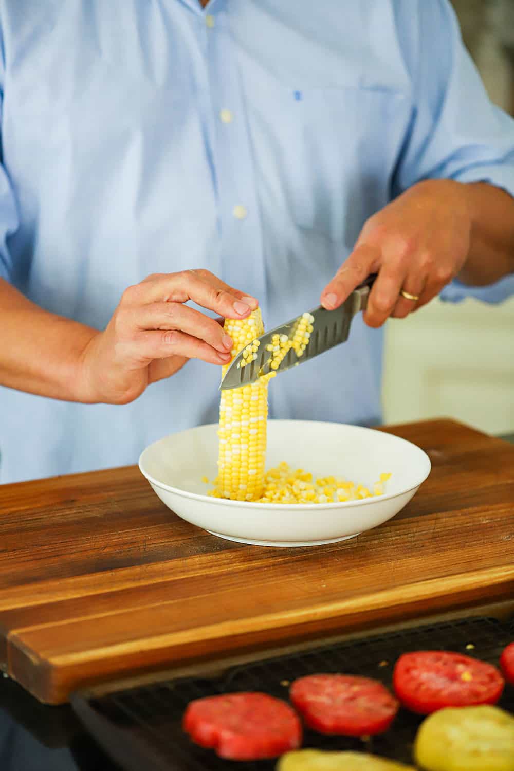 A person holding a ear of fresh corn upright in a bowl and using a knife to cut off the kernels. 