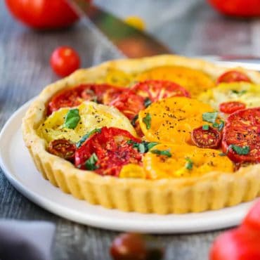 A tomato tart with colorful sliced tomatoes on top in a pastry shell all on a white serving plate.