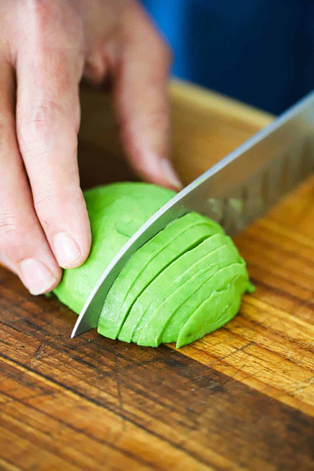 A person holding a peeled, pitted, and halved avocado and using a large knife to cut it into thin slices. 