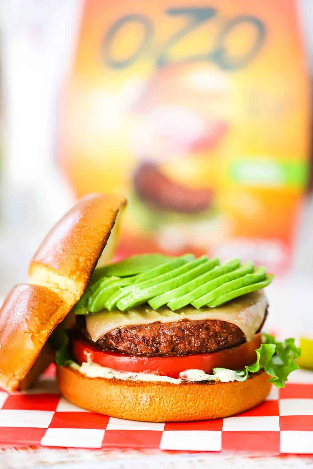 A plant-based California burger topped with sliced avocado sitting on a red-checkered napkin in front of an OZO package. 