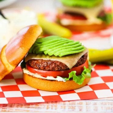 A plant-based California burger topped with sliced avocado sitting on a red-checkered napkin in front of an OZO package.