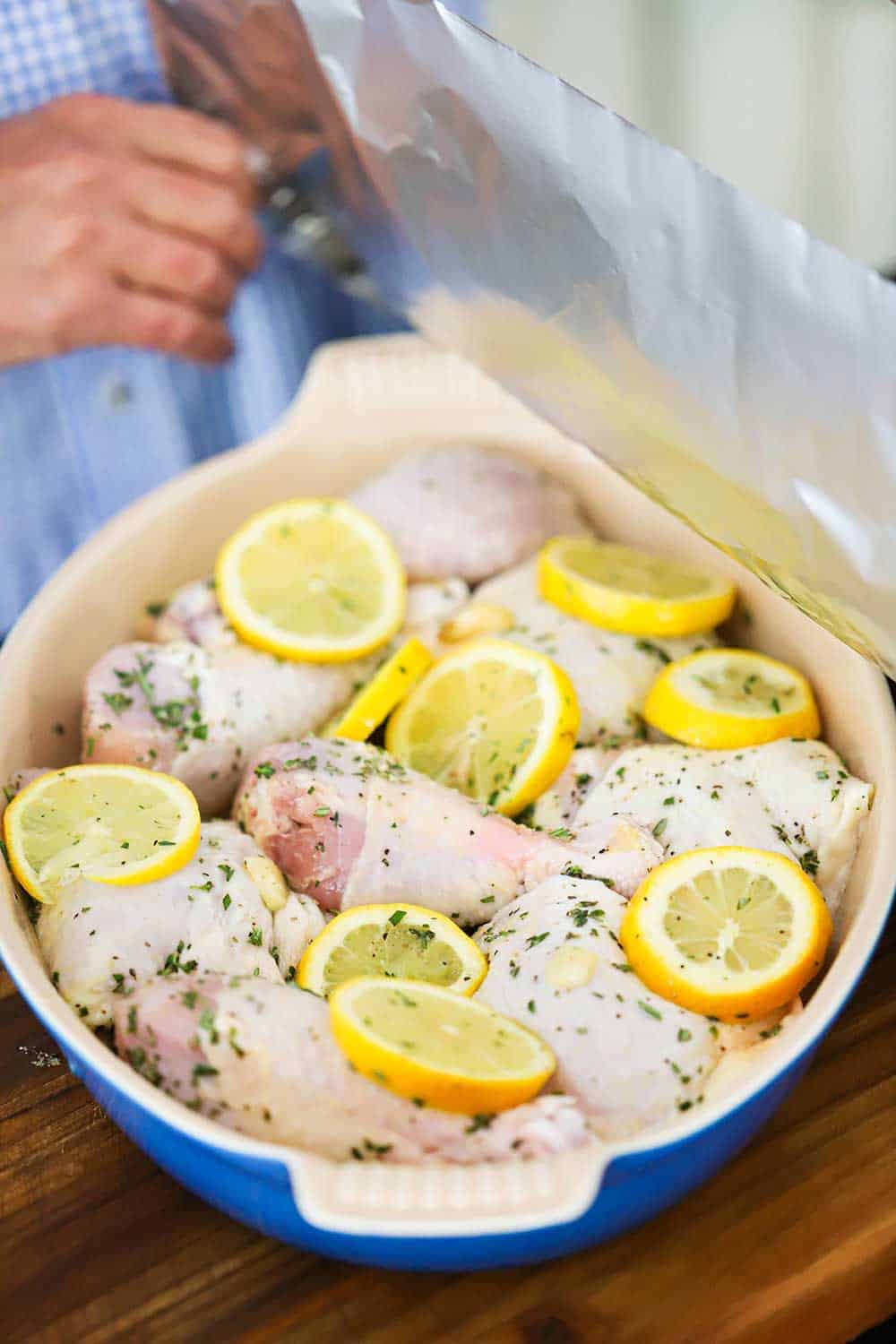 A person placing a piece of foil over a baking dish filled with uncooked chicken pieces, lemon slices, and herbs.