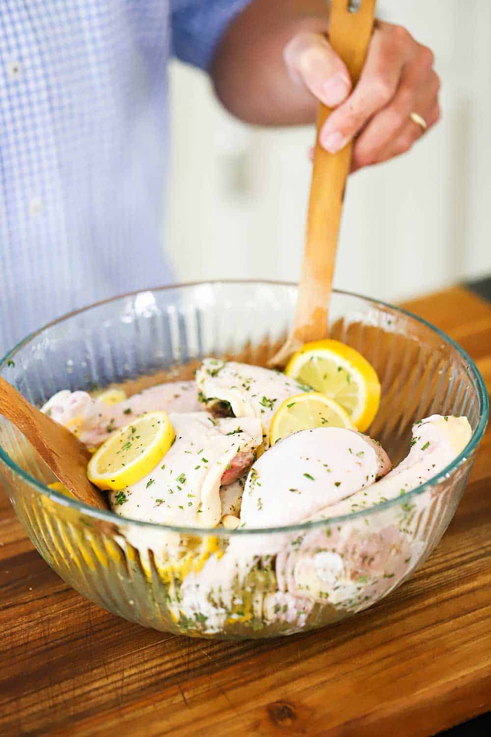 A person using two wooden spoons to mix up uncooked chicken pieces, lemon slices, and herbs in a glass bowl.