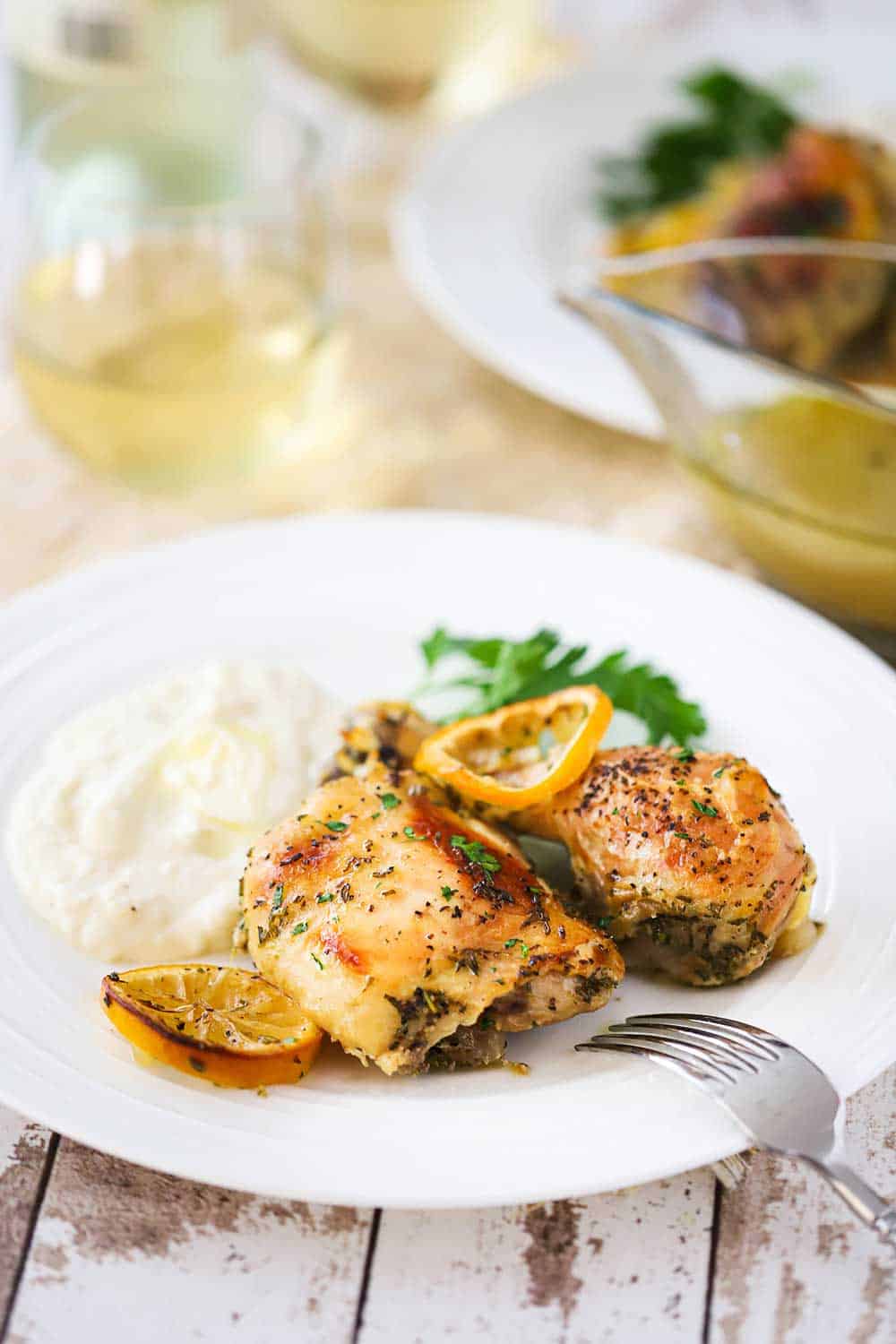 A white plate filled with two roasted chicken pieces, a lemon slice, and mashed potatoes next a glass of white wine.