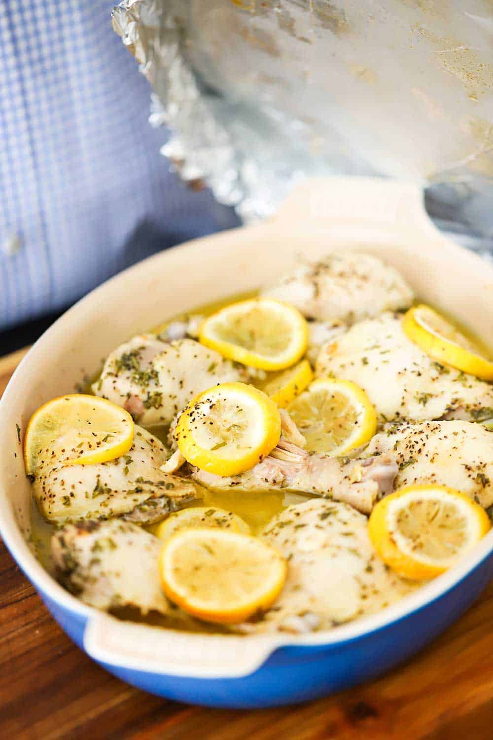 A person removing a piece of foil off a a baking dish filled with cooked chicken with sliced lemons, white wine, and herbs.