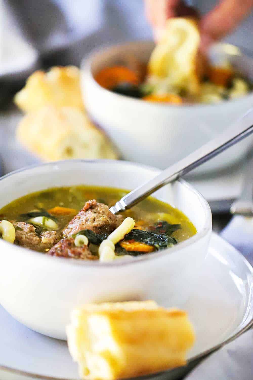 A white soup bowl filled with Italian wedding soup with a spoon in it and piece of bread nearby.